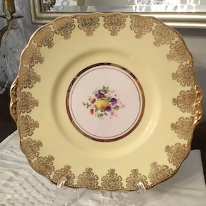 May include: A yellow ceramic serving plate with a gold rim and intricate gold design. The center of the plate features a floral design with fruit.