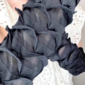May include: A pair of black arm warmers featuring a textured, ruched design with horizontal gathered fabric layers. The sleeves are constructed from a semi-sheer material that creates a repeating pattern of small, raised, petal-like folds along the length.