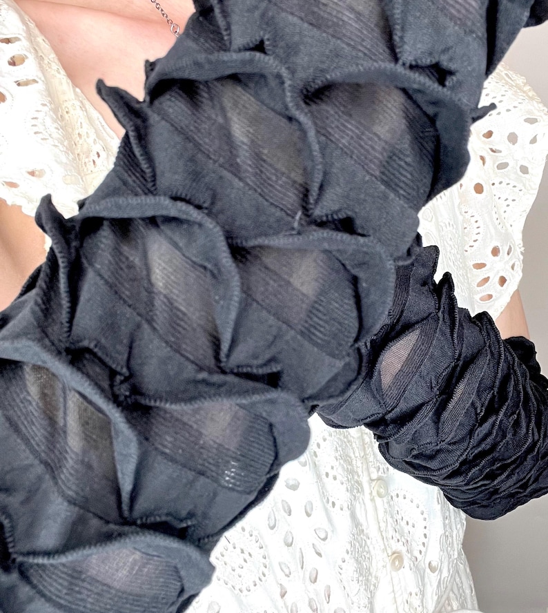 May include: A pair of black arm warmers featuring a textured, ruched design with horizontal gathered fabric layers. The sleeves are constructed from a semi-sheer material that creates a repeating pattern of small, raised, petal-like folds along the length.