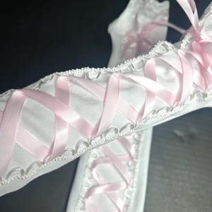 May include: A pair of white arm warmers featuring a crisscross lacing design with light pink satin ribbon. The edges are finished with a white scalloped lace trim, and the fabric has a smooth, solid white appearance throughout the length.