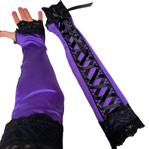 May include: Purple satin arm warmers with black lace trim and a black ribbon corset design. The arm warmers have a fingerless design and are made of a shiny, smooth fabric. The lace trim is at the wrist and upper arm.