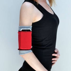 May include: A red, gray, and black arm band with a pocket for a phone. The arm band is worn on the upper arm of a person.