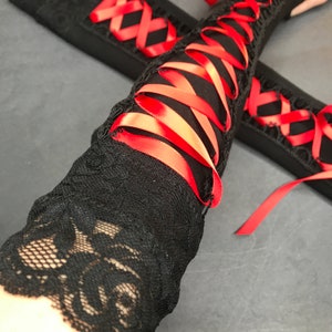 May include: A pair of black arm warmers featuring a lace cuff with a floral pattern and a central section laced with a vibrant red satin ribbon. The design includes a crisscross lacing pattern that runs along the length of the black fabric sleeve.