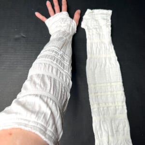 May include: A pair of white arm warmers featuring a textured, ruched, and gathered fabric design. One arm warmer is worn on an arm, extending from the fingers to the upper forearm, while the other is laid flat, showcasing the consistent crinkled pattern.