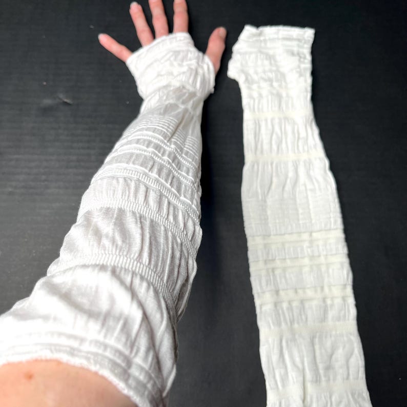 May include: A pair of white arm warmers featuring a textured, ruched, and gathered fabric design. One arm warmer is worn on an arm, extending from the fingers to the upper forearm, while the other is laid flat, showcasing the consistent crinkled pattern.