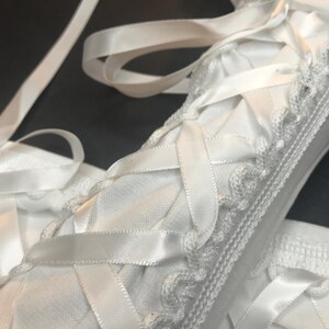 May include: A white fabric item featuring a crisscross lacing pattern made of smooth satin ribbon. The edges are trimmed with a delicate, scalloped crochet border. The material has a fine mesh texture, and long ribbon tails extend from the top.