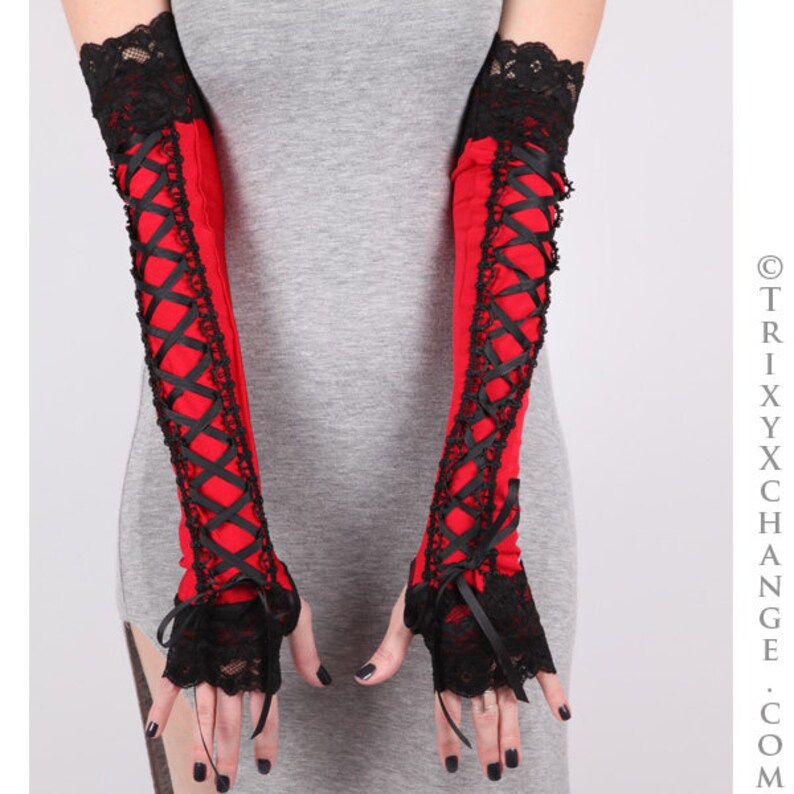 May include: Red and black arm warmers with a gothic aesthetic. The arm warmers feature a red fabric base, black lace trim, and black ribbon criss-cross detailing. The wrist and upper arm cuffs are trimmed with black lace.