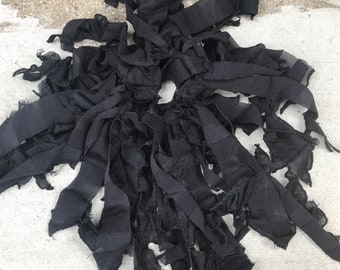 May include: A collection of black fabric strips with frayed, raw edges arranged in a circular, gathered cluster. The strips vary in width and length, featuring a semi-sheer, lightweight texture with visible stitching along the edges of the fabric pieces.