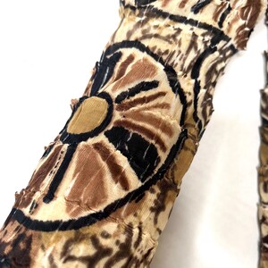 May include: A long, tubular fabric sleeve featuring a crinkled, textured surface. The material displays a repeating pattern of large, circular motifs in shades of tan, dark brown, and black, with solid black outlines defining each circular shape.