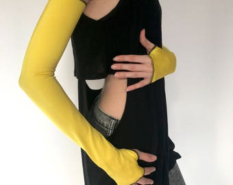 May include: A pair of yellow fingerless gloves with black stitching. The gloves are worn by a person wearing a black top and jeans.