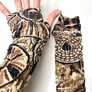 May include: A pair of fingerless gloves featuring a crinkled, semi-sheer fabric with a brown, black, and beige abstract geometric pattern. The gloves have a textured, layered appearance with a thumb hole and extend to cover the forearm area.