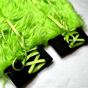 Puede incluir: Primer plano de una tela de piel sintética verde neón con dos puños negros. Los puños están atados con cinta de raso verde brillante. La cinta está atada en lazos. El fondo es blanco.