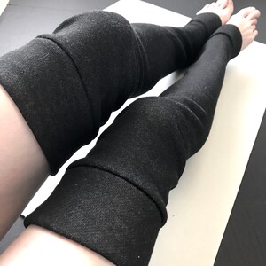 Puede incluir: Calentadores de piernas negros hechos de una tela suave y texturizada. Los calentadores de piernas se muestran desde las rodillas hacia abajo, con los pies visibles. La tela es de color carb&oacute;n oscuro.