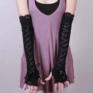 May include: A pair of black fingerless arm warmers featuring a lace trim at the top and bottom edges. The sleeves are designed with a crisscross ribbon lacing detail running down the center, finished with a small bow at the wrist area.