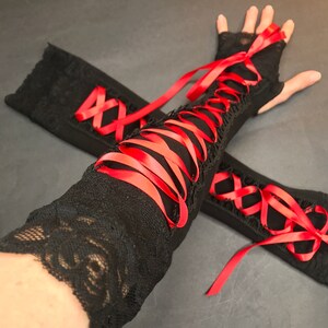 May include: A pair of black fingerless gloves featuring intricate lace detailing and a vibrant red ribbon corset-style lacing that runs along the length of the forearm. The gloves are constructed from a soft fabric with lace trim at the edges.