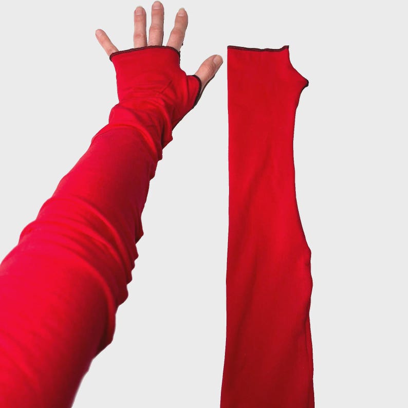 May include: A pair of bright red fingerless arm warmers featuring a long, fitted silhouette that extends past the elbow. Each sleeve includes a dedicated thumb hole and a thin, dark-colored trim along the top edge, providing a clean finish.