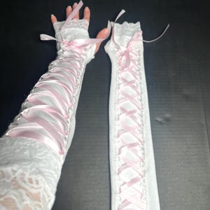 May include: A pair of white fingerless gloves featuring a lace trim at the top and bottom edges. The gloves are decorated with a light pink satin ribbon laced in a crisscross pattern down the center, tied into a bow at the top of each glove.