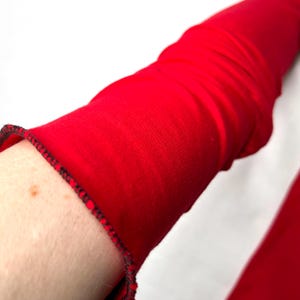 May include: A vibrant red long-sleeved garment featuring a fitted silhouette and a distinct lettuce-edge hemline. The cuff is finished with a delicate, dark-colored serged stitch that provides a subtle contrast against the bright red fabric.