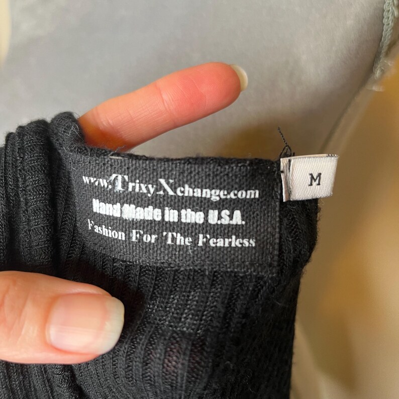 May include: Close-up of a black ribbed fabric with a tag. The tag reads "www.TrixyXchange.com Hand Made in the U.S.A. Fashion For The Fearless" and has a size "M" tag attached.