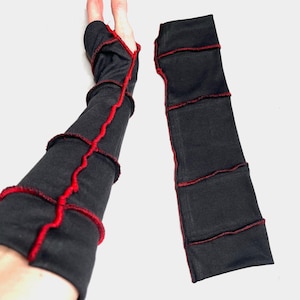 May include: A pair of black fingerless gloves with red stitching. The gloves are made of a dark fabric and feature a patchwork design with visible red thread outlining the seams and edges. The gloves are designed to cover the forearm and hand.
