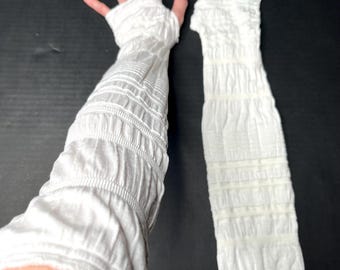 May include: A pair of white arm warmers featuring a textured, ruched, and gathered fabric design. One arm warmer is worn on an arm, extending from the fingers to the upper forearm, while the other is laid flat, showcasing the consistent crinkled pattern.