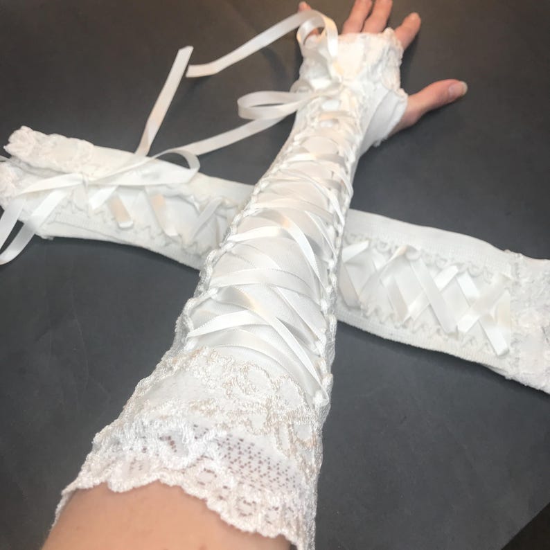 May include: A pair of white fingerless gloves featuring intricate lace trim at the top and bottom edges. The gloves are designed with a corset-style lace-up closure using white satin ribbon that runs along the length of the forearm.