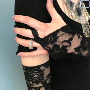 May include: A black lace fingerless glove with a ring on the index finger. The glove is worn on the left hand and the ring is a silver band with a clear stone.