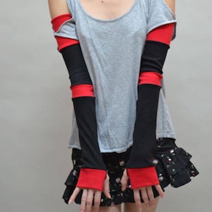 May include: Gray tank top with cut-out shoulders, paired with black and red arm warmers. The arm warmers have a black base with red cuffs and a red band at the upper arm. A black floral skirt completes the outfit.