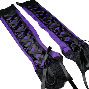 May include: A pair of purple leg warmers with black ribbon criss-cross detailing and black lace trim. The leg warmers have black satin bows at the top and bottom. The background is white.