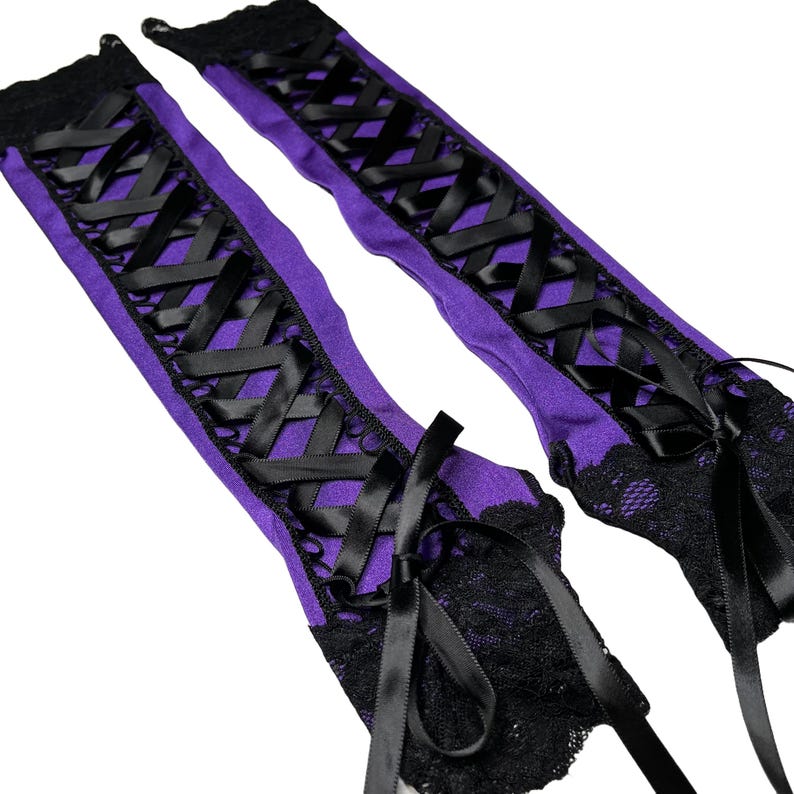 May include: A pair of purple leg warmers with black ribbon criss-cross detailing and black lace trim. The leg warmers have black satin bows at the top and bottom. The background is white.