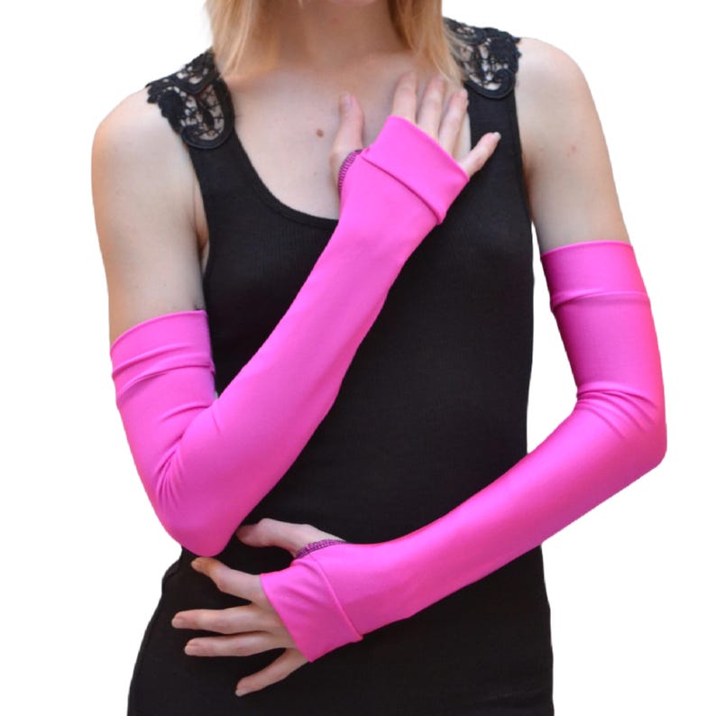 May include: A pair of long pink arm warmers made of a stretchy, shiny fabric. The arm warmers have a thumb hole and a folded cuff at the wrist.