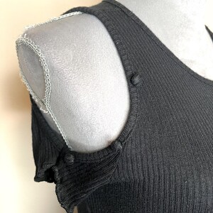 May include: Close-up of a black ribbed knit top with a scoop neckline and shoulder cutouts. The top features small buttons at the shoulder seams and a decorative trim along the shoulder line. The fabric has a vertical ribbed texture.