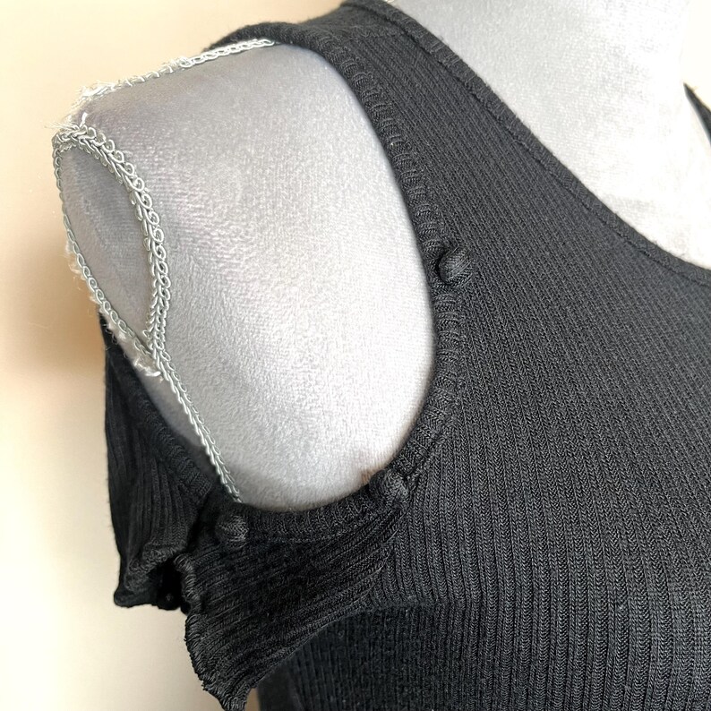 May include: Close-up of a black ribbed knit top with a scoop neckline and shoulder cutouts. The top features small buttons at the shoulder seams and a decorative trim along the shoulder line. The fabric has a vertical ribbed texture.