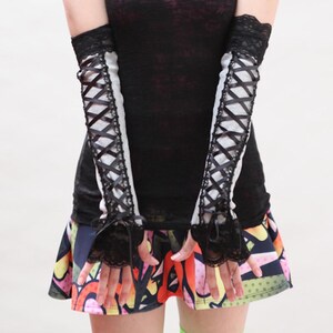 May include: A pair of light gray arm warmers featuring black lace trim at the top and bottom edges, with a black ribbon crisscross lacing design running down the center of each sleeve, finished with small black ribbon bows at the wrists.