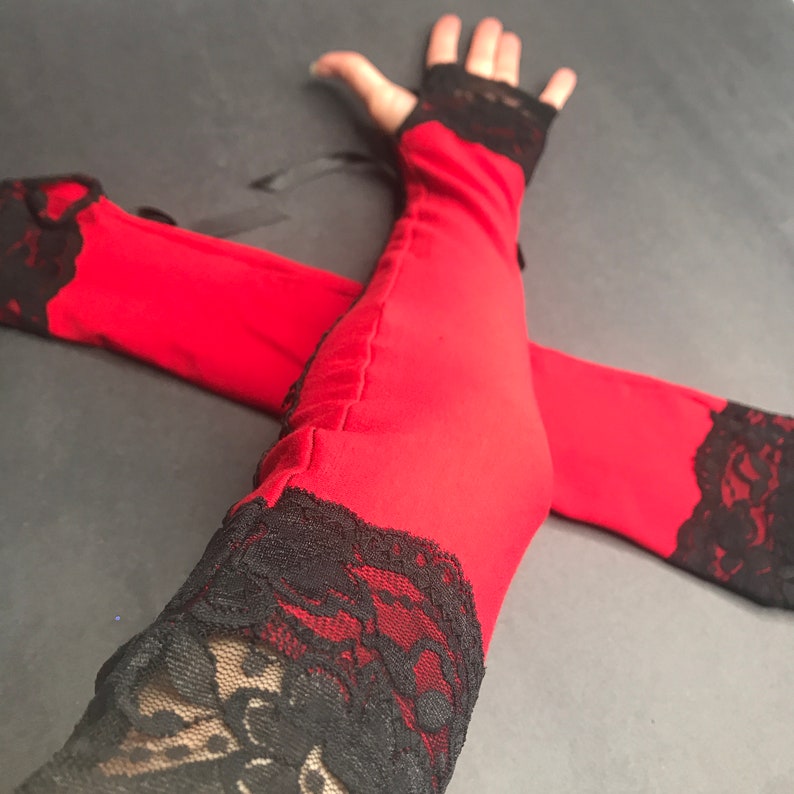 May include: A pair of fingerless arm warmers in bright red fabric featuring black lace trim at the top and bottom edges. The arm warmers include thin black ribbon ties at the upper section, providing a secure fit around the arm with a visible seam.
