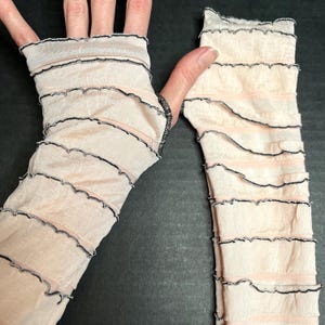 May include: A pair of fingerless arm warmers made from a sheer, pale peach-colored fabric. The sleeves feature horizontal, ruffled layers with delicate, dark-colored lettuce-edge stitching along the borders of each tier, creating a textured look.