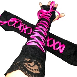 May include: A pair of black fingerless gloves featuring intricate lace detailing and a vibrant hot pink ribbon laced in a crisscross pattern up the forearm. The gloves extend past the wrist, providing a decorative aesthetic with contrasting colors.