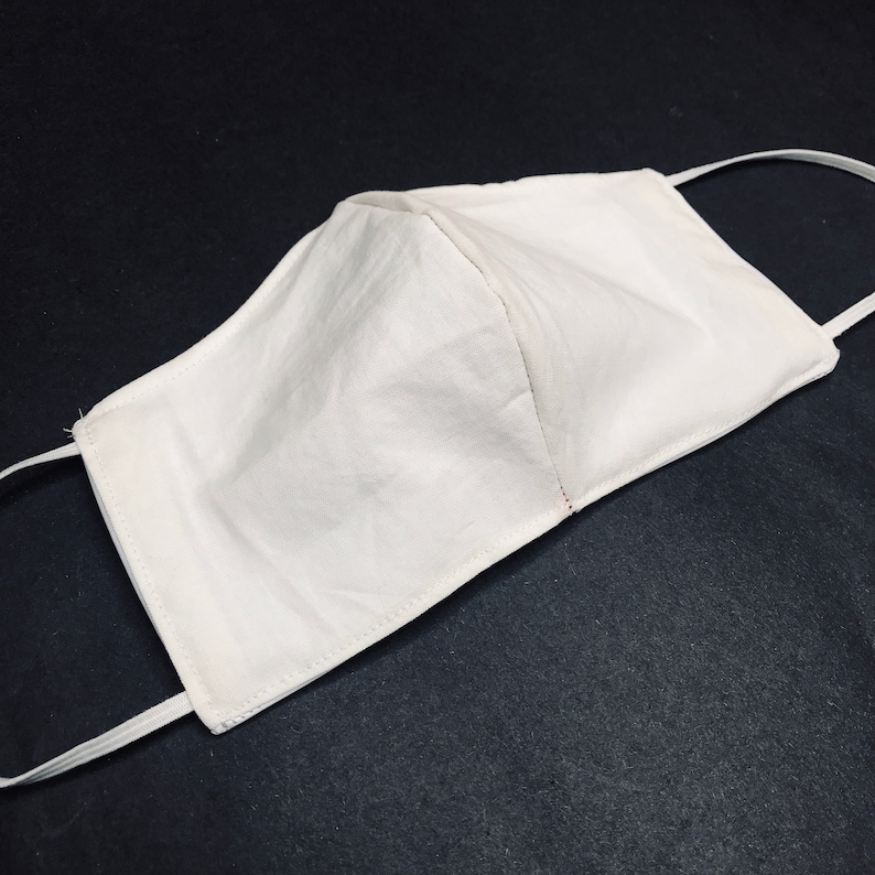 May include: A plain white fabric face mask featuring a structured center seam and thin white elastic ear loops. The mask has a rectangular shape with tapered sides and visible stitching along the edges, providing a simple and functional design.