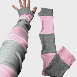 Puede incluir: Calentadores de brazo a rayas grises y rosas con agujeros para los pulgares. Los calentadores de brazo están hechos de un material suave y presentan un puño gris y rayas rosas y grises. Los calentadores de brazo se muestran con una mano.