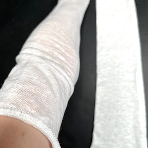 May include: Close-up of a white, sheer sleeve made of a lightweight, textured fabric. The sleeve is slightly gathered, revealing the skin underneath. The fabric has a subtle, natural texture, and the cuff is finished with a simple hem.