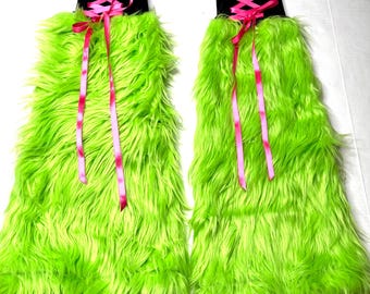 May include: Two neon green faux fur leg warmers with black tops and pink ribbon accents. The leg warmers have a long, shaggy texture and are designed to be worn on the legs. The pink ribbon is tied in a criss-cross pattern.