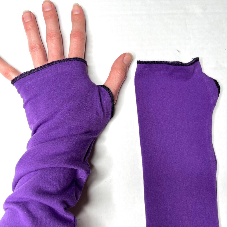 May include: A pair of vibrant purple fingerless gloves made of a smooth, stretchy fabric. Each glove features a dedicated opening for the thumb and a finished black stitched edge along the top and thumb hole, providing a snug fit for the arm.