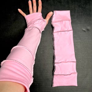 May include: Pink fingerless gloves with black stitching. The gloves are made of a soft material and have a patchwork design. One glove is on a person's hand, and the other is lying flat.