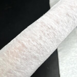 May include: Close-up of a rolled-up, off-white fabric. The material appears to be a loosely woven linen or cotton, with a slightly textured surface. The fabric is draped over a dark surface.