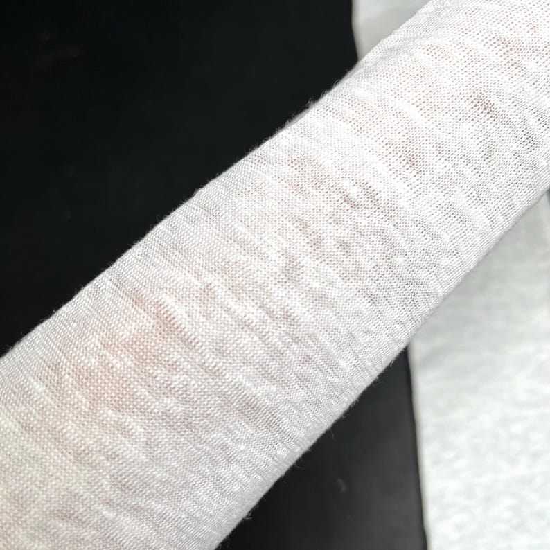 May include: Close-up of a rolled-up, off-white fabric. The material appears to be a loosely woven linen or cotton, with a slightly textured surface. The fabric is draped over a dark surface.