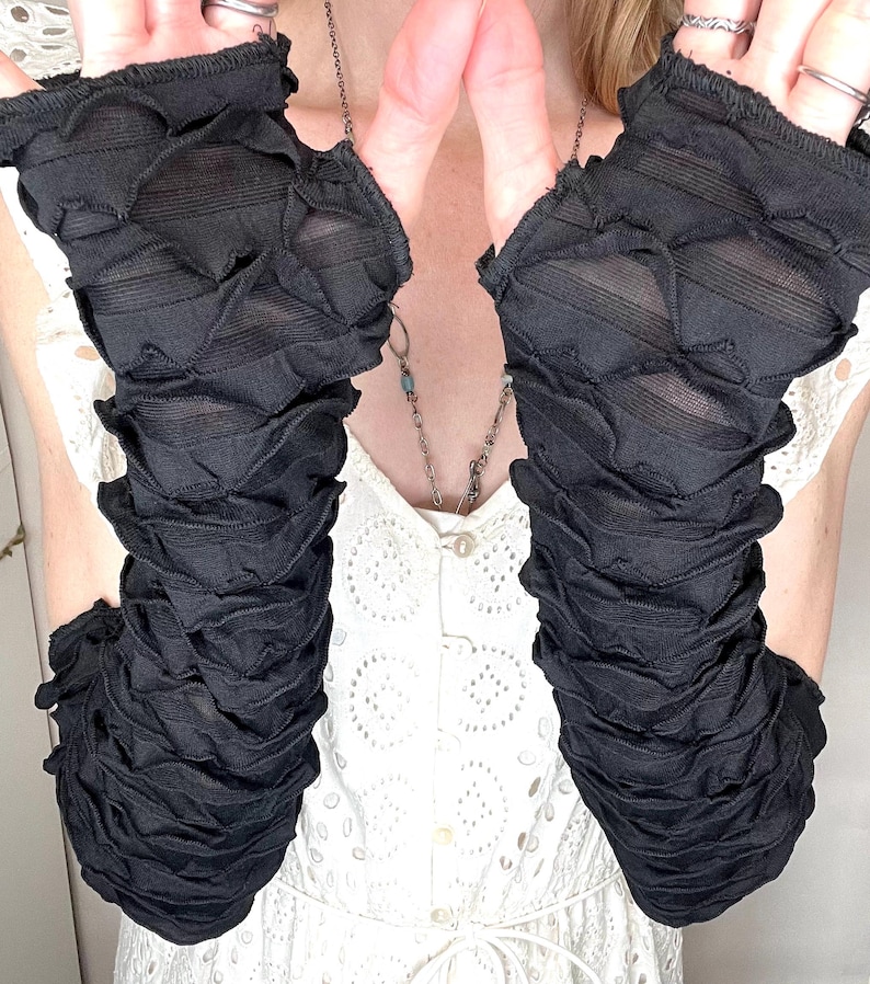 May include: A pair of black fingerless gloves featuring a textured, ruched, and layered fabric design. The gloves extend from the knuckles to the mid-forearm, showcasing a repeating horizontal pattern of gathered material throughout the length.