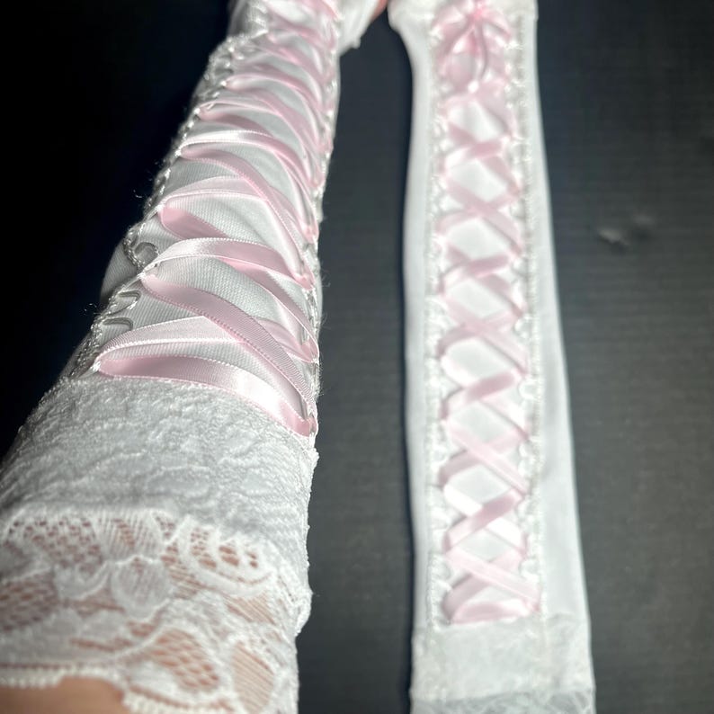 May include: A pair of long white gloves featuring a lace cuff at the wrist and a corset-style lace-up design running the length of the forearm. The lacing is composed of a smooth, light pink ribbon threaded through white fabric eyelets.