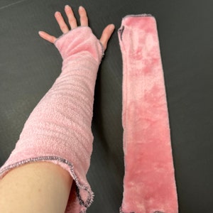 May include: Pink fingerless gloves with a soft, plush texture. The gloves have a ruffled edge and a gray trim. One glove is on a person's arm, and the other is lying flat.