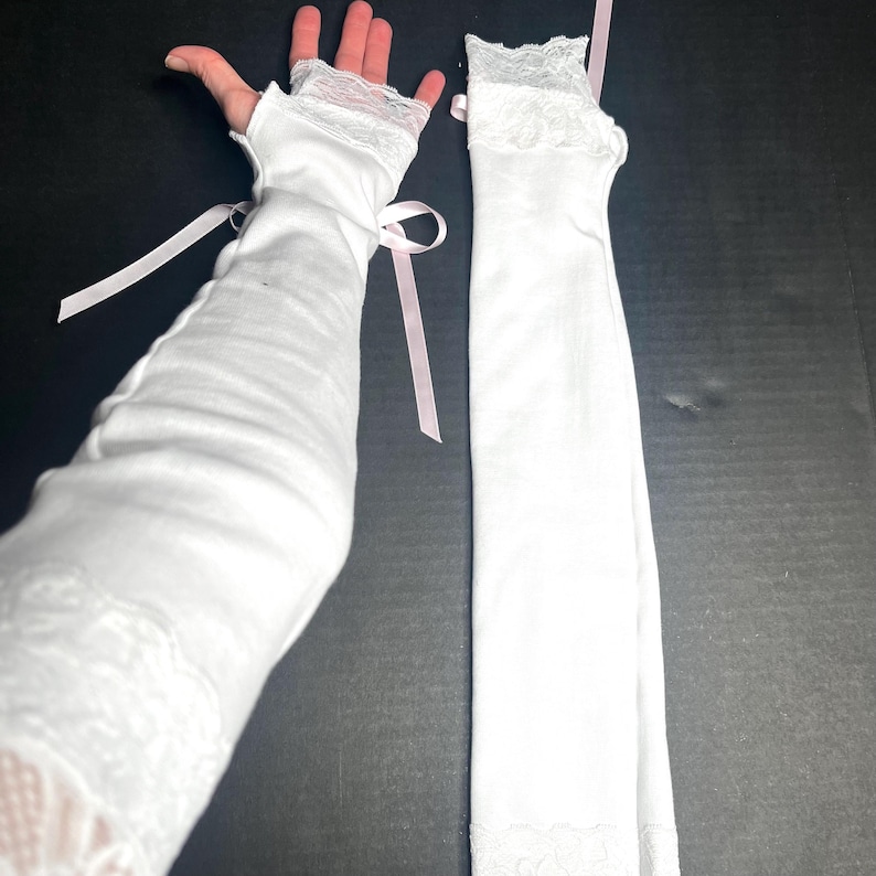 May include: A pair of white fingerless arm warmers featuring delicate lace trim at the top and bottom edges. The fabric is smooth and opaque, accented with thin, light pink ribbon ties that are tied into bows on the sides of the arm warmers.