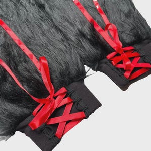 May include: A pair of black faux fur leg warmers featuring a long-pile texture and a black ribbed knit cuff. Each leg warmer is decorated with a vibrant red satin ribbon laced through black loops in a crisscross pattern, tied into a bow.
